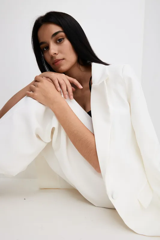 Young woman in white suit sitting thoughtfully against minimalistic white background, showcasing modern fashion style and calm mood in a studio setting.