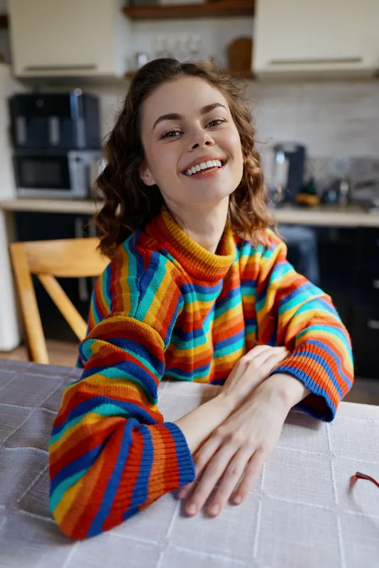 Smiling woman with rainbow striped sweater sits at a kitchen table, enjoying a cozy home moment, casual lifestyle, and friendly atmosphere, with natural light and warm colors in a bright kitchen