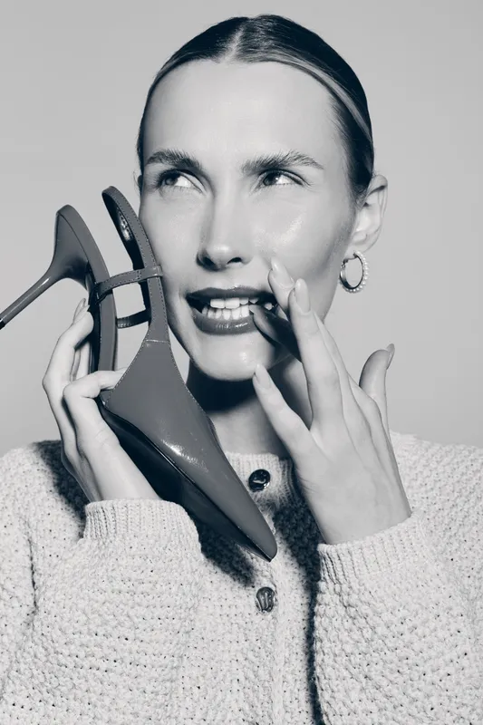 Caucasian woman with fair skin holds red high heel shoe near face, wearing knitted sweater and hoop earrings. Fashion concept with minimalistic studio lighting and neutral background.
