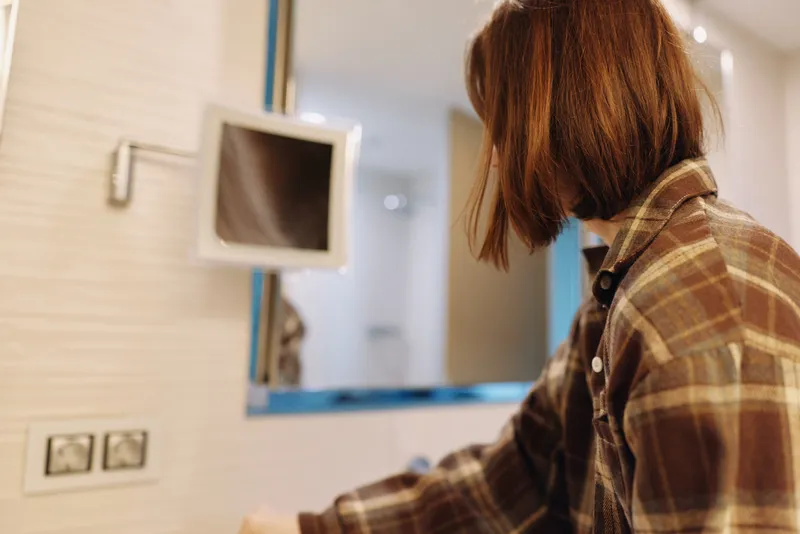 Person in plaid shirt examining a mirror in a minimalist hotel room, soft lighting, desk and outlets, capturing a calm, thoughtful moment.