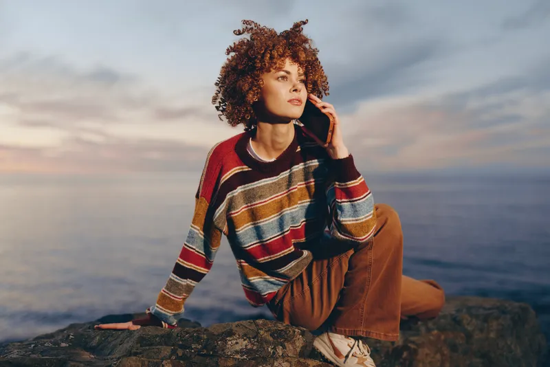 Woman lifestyle portrait outdoors, curly haired woman in striped sweater sits on rocky cliff, ocean waves behind her, hand on hair, relaxed mood at sunset, natural soft light