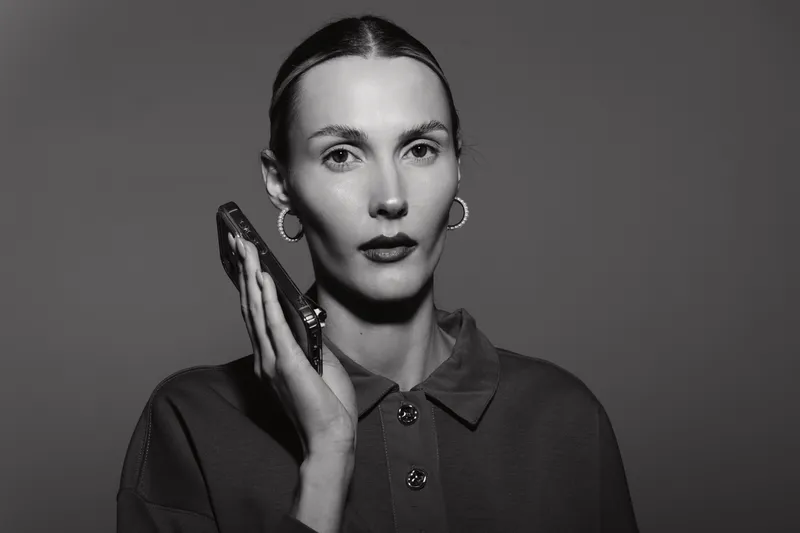 Portrait of a pale white woman with dark hair slicked back, holding a smartphone to her ear. She wears a dark top and hoop earrings in a monochrome studio. Concept of communication and focus.