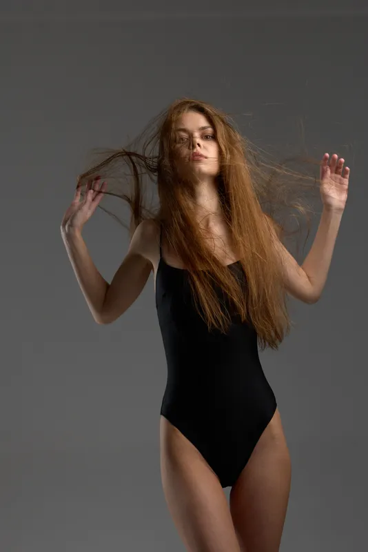 young woman in black bodysuit with long hair posing confidently on gray background