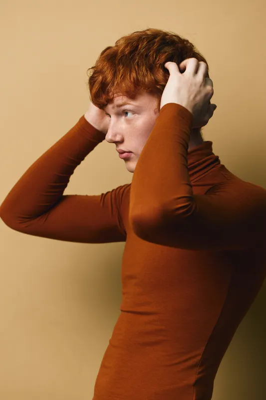 Redhead man with curly hair wearing brown turtleneck poses with hands in hair, looking sideways on beige background in studio portrait with soft natural light.