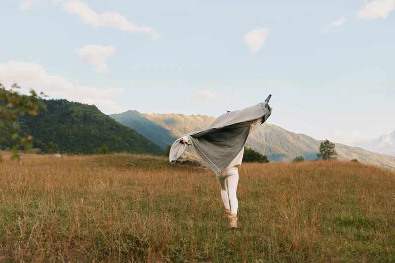 A woman in a light shawl stretches in a golden meadow with distant mountains, conveying freedom and calm in the open countryside, an airy scene of movement under a bright sky.