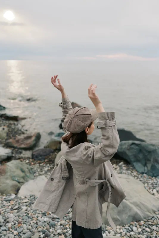 A woman in a beige jacket and hat stands on a rocky beach with arms raised, facing the calm sea under a cloudy sky. Concept of freedom, nature, and peaceful moments.