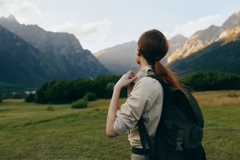 Woman with a backpack stands in a wide valley, gazing toward distant mountains, embracing outdoor exploration and nature. Soft natural light enhances the calm mood.