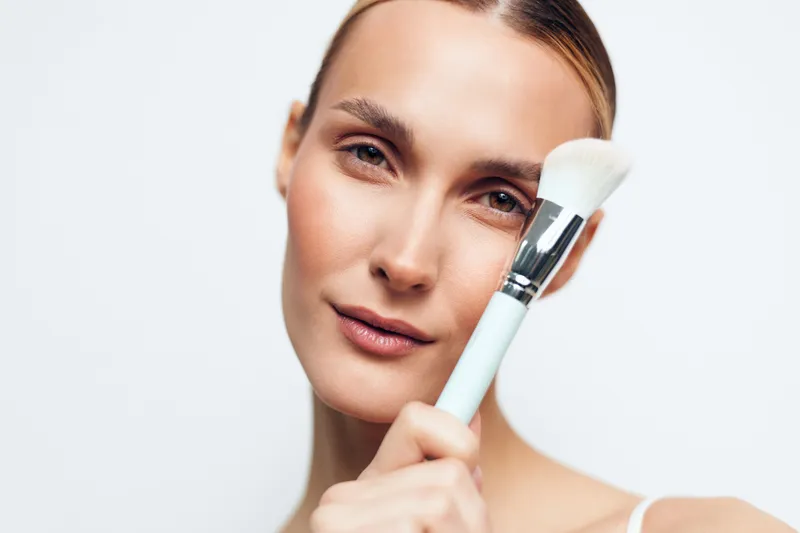 Young woman applying makeup with a brush on her face, closeup portrait, natural skin, soft lighting, white background, beauty routine, cosmetic application, healthy complexion