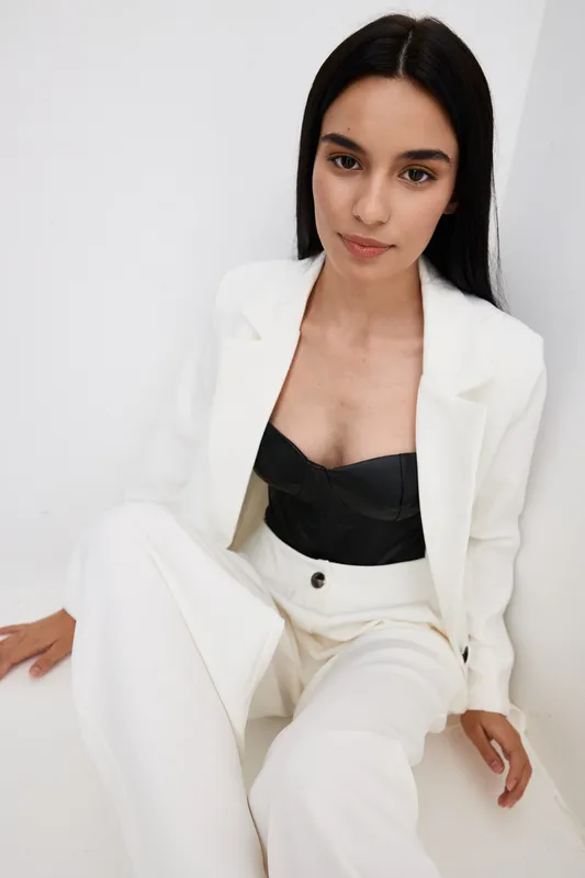 Young woman wearing white suit and black top seated against white background, showing natural makeup, long dark hair, and confident expression in minimalist style.