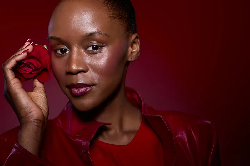 Close-up portrait of an elegant black woman with dark skin holding a red rose near her face, wearing a red leather jacket on a dark red background. Concept of beauty and confidence.