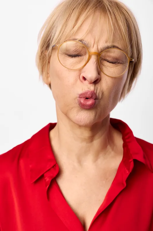 Woman with short blonde hair and glasses applying white face cream, eyes closed, closeup on clean white background, red shirt, skincare wellness selfcare, soft studio light
