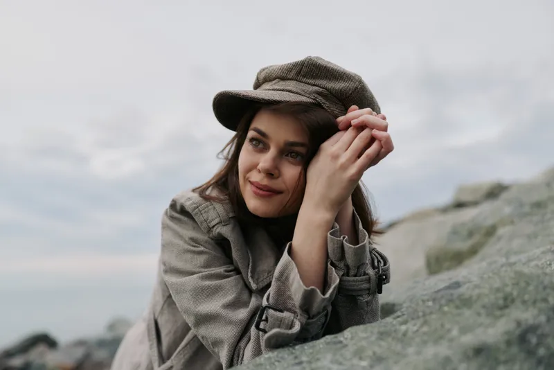 Young woman in casual coat and cap rests on rock near sea, smiling softly. Overcast sky and natural coastal background create calm, contemplative outdoor mood and lifestyle concept.