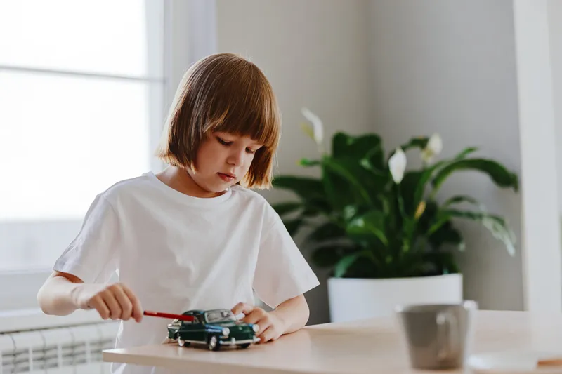 A young boy playing with a toy car, focusing intently while using a red tool. The bright indoor setting is lively and inviting, creating a playful atmosphere.