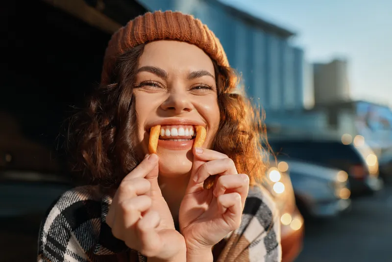 Smiling woman in a beanie holds a snack to her teeth in a playful candid urban portrait with golden hour glow, authenticity and emotional storytelling for mindful living lifestyle.