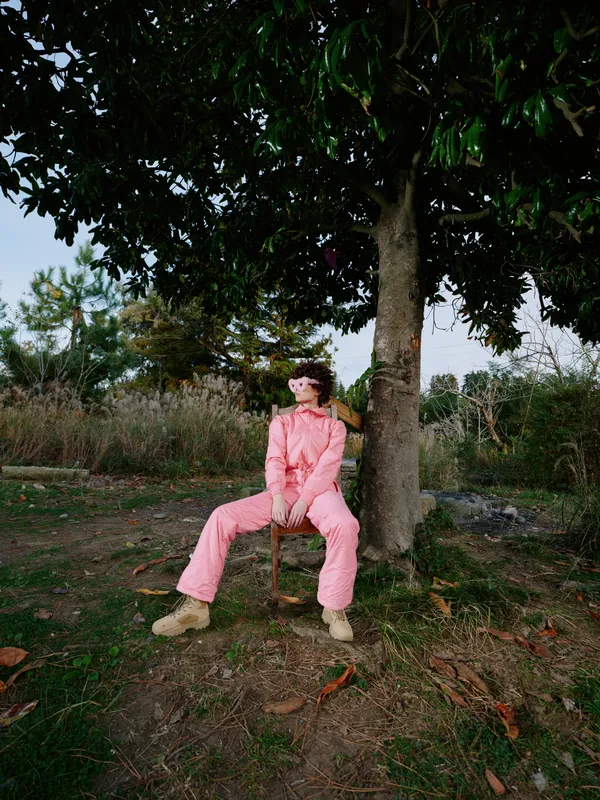 Outdoor portrait of a woman sitting under a tree in a pink jumpsuit, relaxed pose in a natural setting with daylight, casual style among leaves and grass, warm tones.