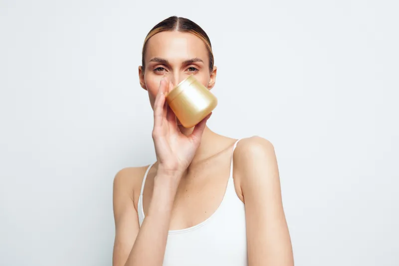 Young woman with clear skin holding golden jar near face on white background. Concept of skincare, beauty, wellness, and self-care with natural light and minimalistic style.