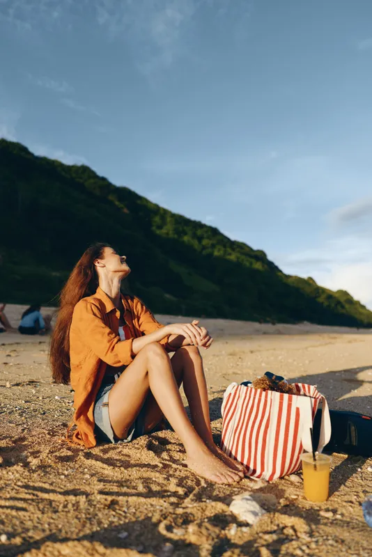 Woman beach sand seaside summer relax striped sunlight, casual portrait sitting on rocky shore, striped bag on sand, straw hat and scarf, calm vacation by blue sky