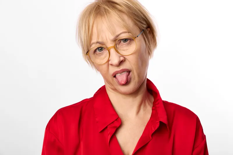 Playful older woman portrait with blonde hair and glasses, tongue out, wearing red shirt, looking at camera, studio shot on plain white background, closeup