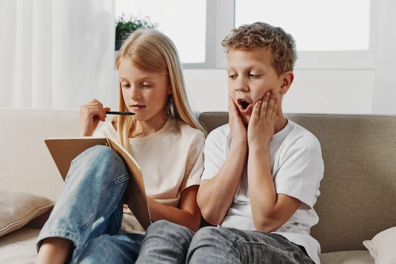 Curious boy and focused girl sitting on couch playing a guessing game with pencil and paper in bright living room. Concept of childhood fun, learning, and communication.