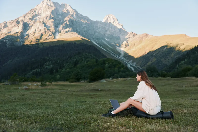 A solitary person sits on a grassy field with a laptop, set against a scenic mountain range. The calm outdoor setting communicates productivity, focus, and a peaceful work mindset.