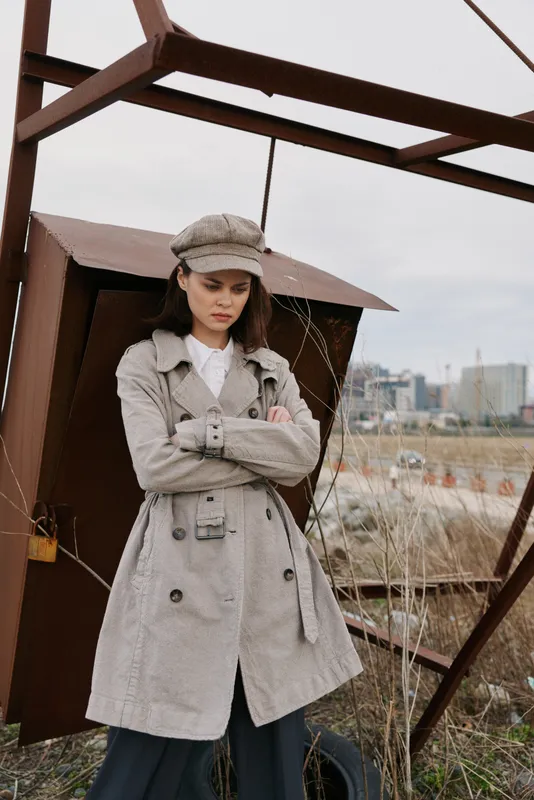 Young woman in long coat and hat stands with crossed arms near rusty metal structure outdoors. Urban background with dry grass and industrial elements visible. Fashion concept outdoors.