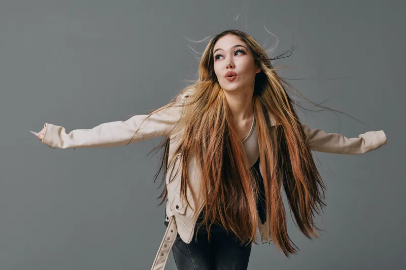 Young woman with long hair wearing light jacket making playful face with arms stretched out on gray background in studio. Fashion, beauty and fun portrait.