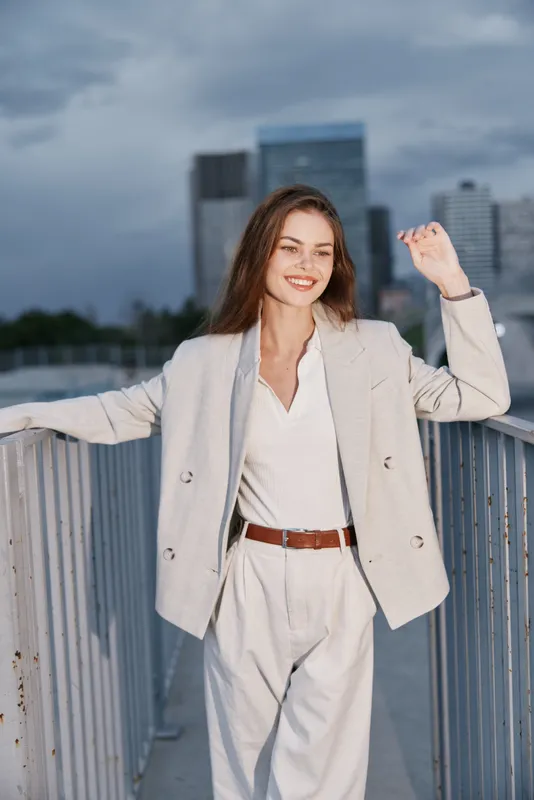 Young confident woman in light business suit smiling outdoors with urban skyline in background, showcasing professional fashion and positive energy