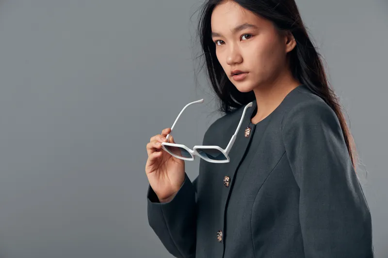 Young woman with long dark hair holding white sunglasses wearing dark gray jacket looking to side on gray background. Beauty, fashion and casual portrait with serious expression and confident pose.