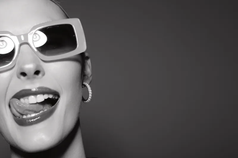 Closeup of a smiling white woman wearing sunglasses and earrings with tongue out. Studio portrait with playful and fun mood.