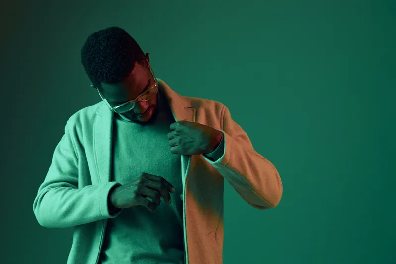 Stylish African man adjusting his coat against a vibrant green background, showcasing modern fashion and confidence in a contemporary setting