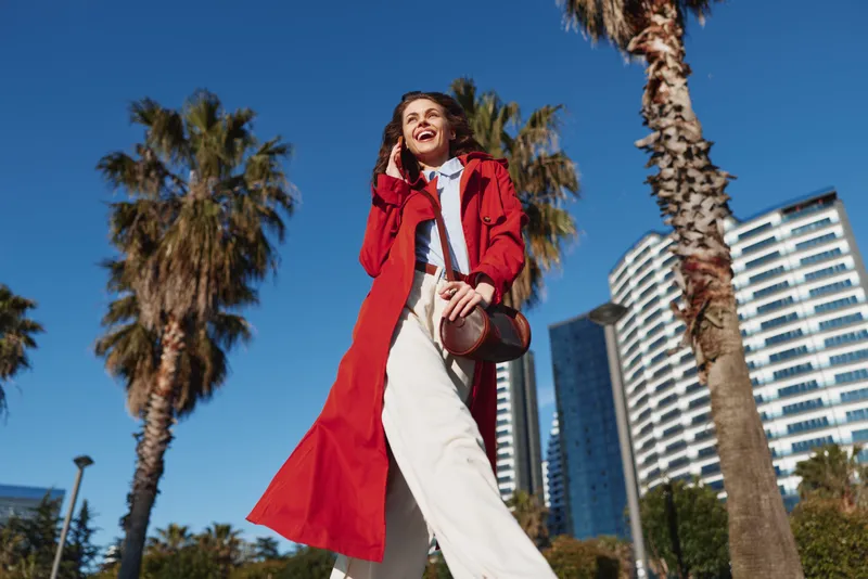 Happy woman in red coat walking outdoors near palm trees and modern buildings under clear blue sky. Concept of urban lifestyle and casual fashion.
