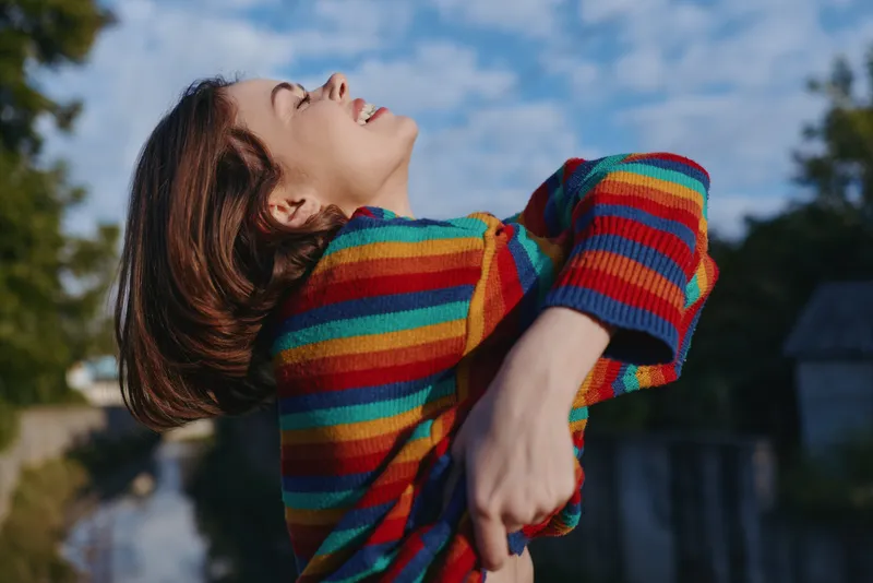 Woman in a rainbow striped sweater outdoors by a canal, smiling and joyful young adult leaning back with eyes closed, casual outfit, lifestyle portrait and travel mood.