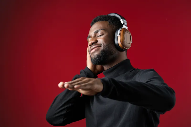 Young African man enjoying music with headphones on a red background, expressing joy and relaxation in casual black attire