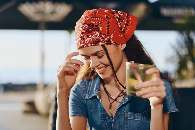 Lifestyle woman with modern boho-western denim street style smiling happily while holding a drink, relaxed outdoor setting, film color tone enhancing natural mood.