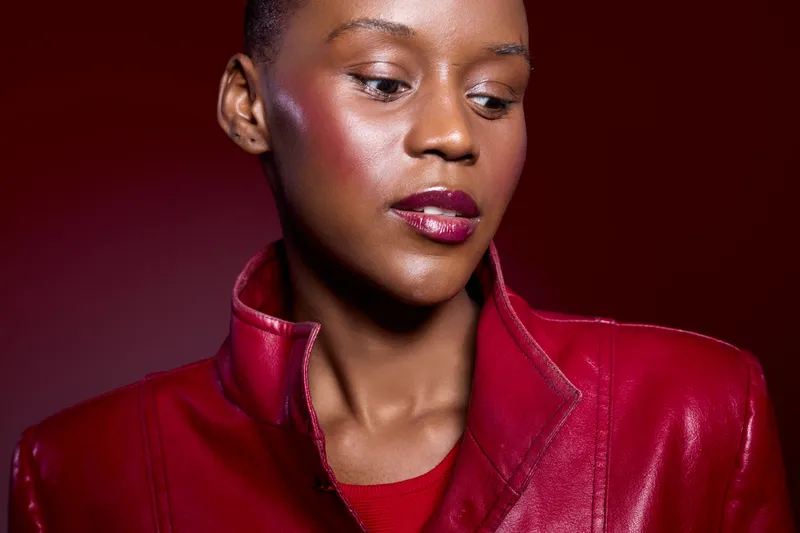 Portrait of a dark skinned African woman wearing a red leather jacket looking down with a calm expression. Studio shot with red background and glossy makeup for fashion concept.