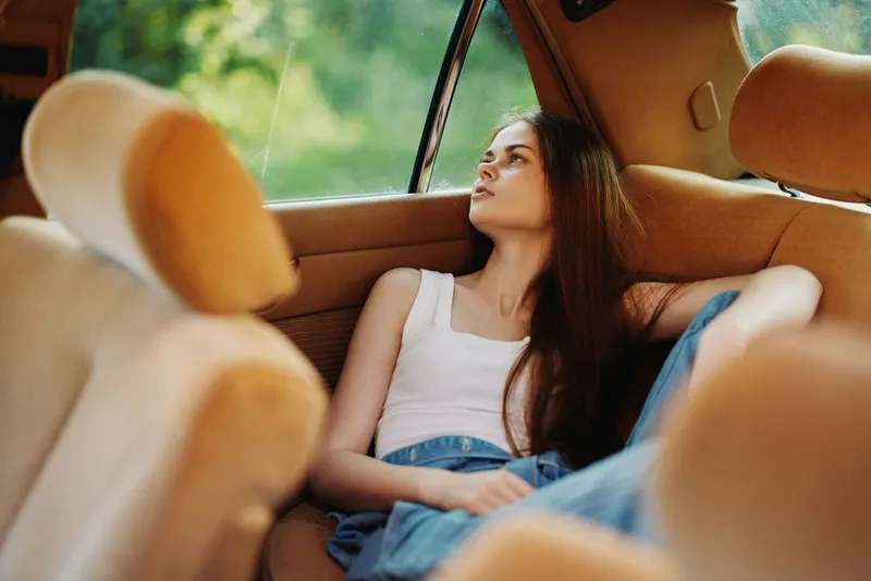 Pensive young woman in a vintage car, reflecting on life through the window, dressed casually in a tank top and jeans, surrounded by warm colors and natural light.