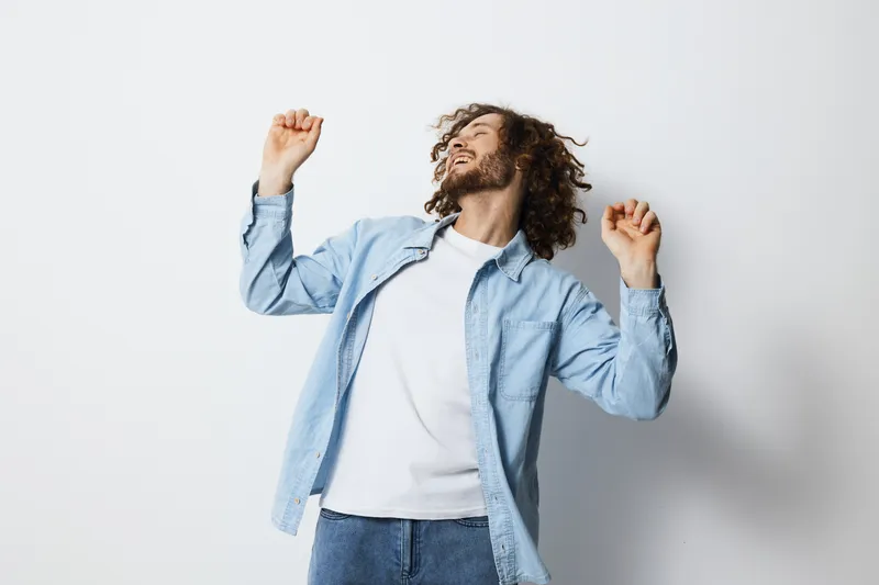 Happy young man with curly hair dancing joyfully in a casual outfit against a light background, embodying carefree emotions and a vibrant lifestyle