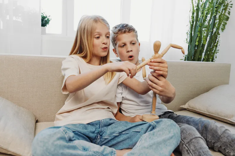 Two children sitting on sofa playing with wooden artist mannequin, exploring movement and creativity in bright living room environment.