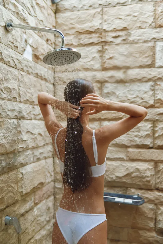 woman taking shower with water falling on her back in rustic stone outdoor bathroom, relaxed mood, natural lighting, summer wellness ritual, healthy lifestyle