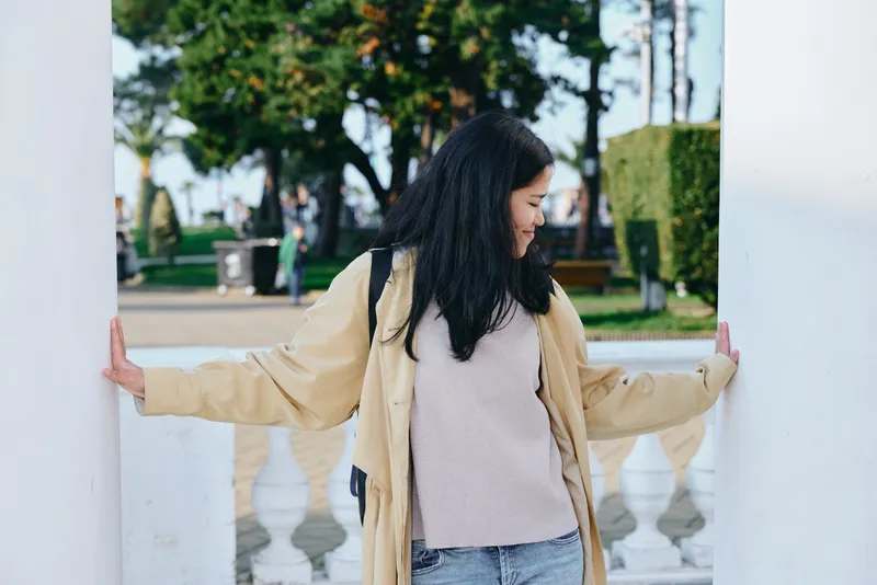 Woman in a beige coat stands between white pillars in a park, arms resting on columns, looking aside with a calm casual mood, outdoor setting with trees and light breeze