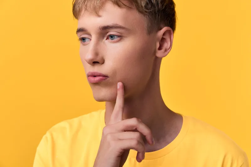 Thinking, contemplation, thoughtful, pondering, finger, chin, male with yellow background, close up studio shot of a young adult contemplating an idea, calm expression, minimal styling, thoughtful