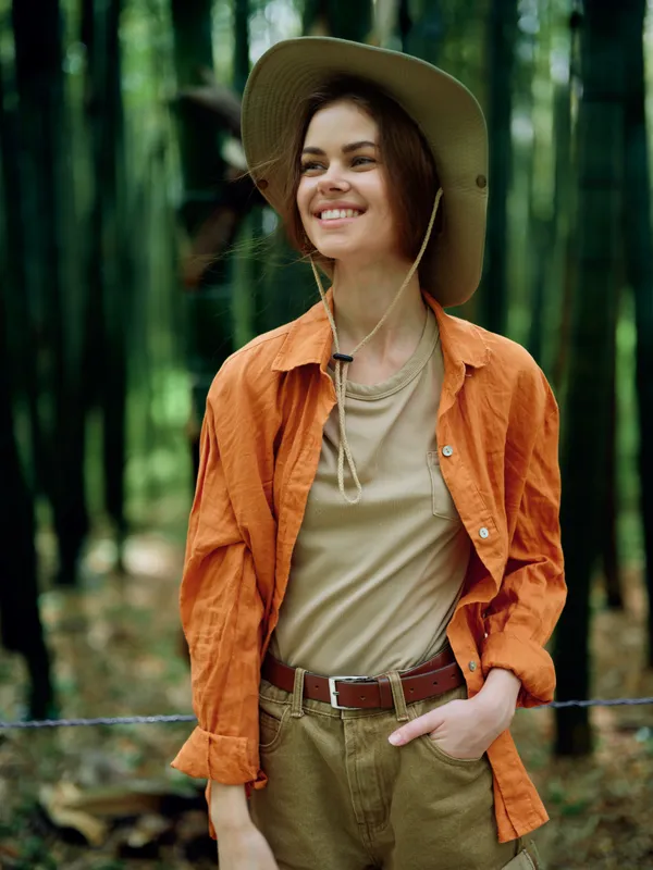 Woman smile hat outdoors forest portrait casual happy young adult in orange shirt and khaki pants, hands in pocket, natural light and relaxed expression during nature walk.