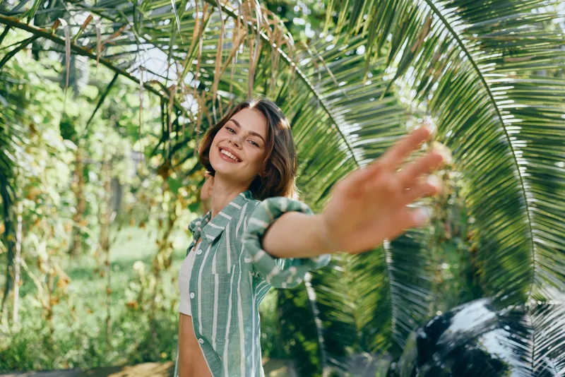 A cheerful woman poses in a lush tropical garden, reaching toward the camera with a sunny smile, wearing a striped green dress, creating a vibrant portrait full of energy and warmth.