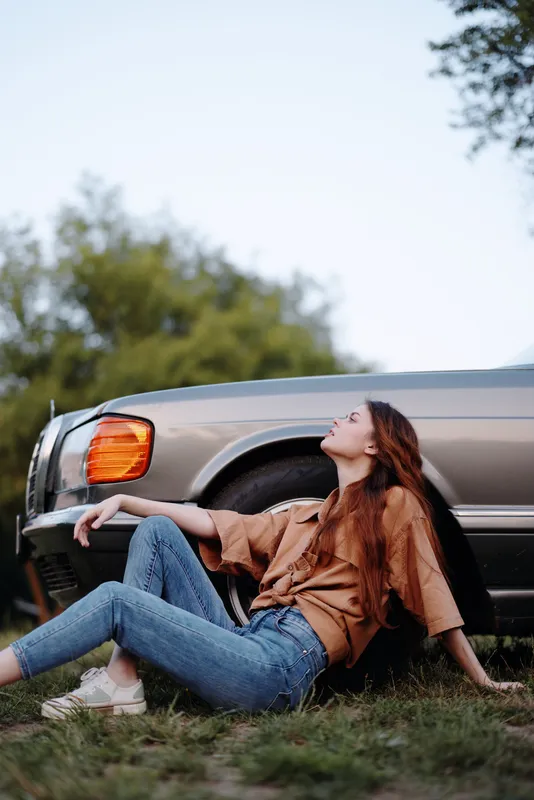 Young woman relaxing outdoors by a vintage car, wearing a casual outfit and exuding a carefree vibe in nature, showcasing fashion and relaxation.