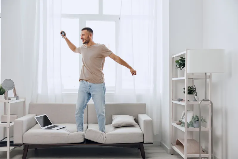 Happy man enjoying music while dancing on the couch in a bright living room, surrounded by plants and modern decor, embodying a lively atmosphere of joy.