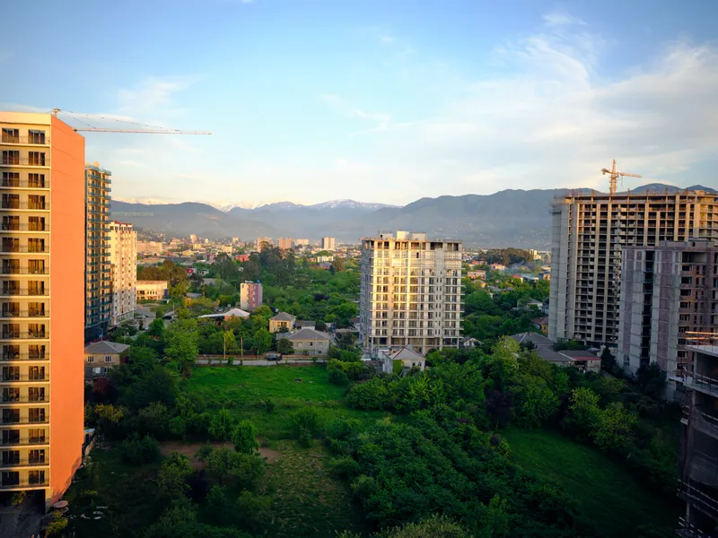 Construction buildings and cranes in city with greenery and skyline view, mountains in background and apartment towers under development, urban landscape at golden hour.