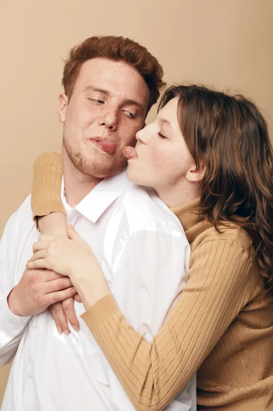 Young couple in casual clothes embraces tenderly, woman kisses man on cheek. Intimate moment of affection and love captured in warm neutral studio setting.