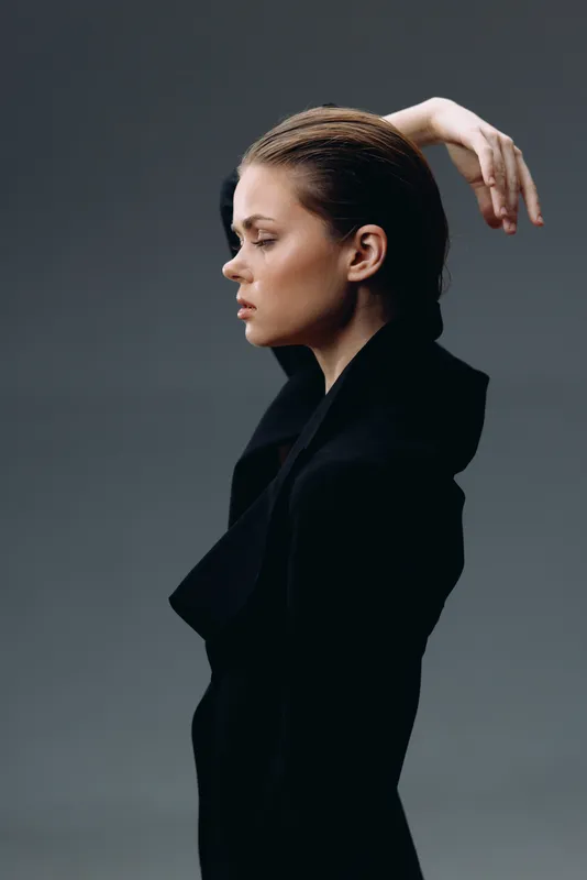 Profile of a serious woman in black coat with hand raised, moody lighting, elegant style, studio background, contemplative mood.