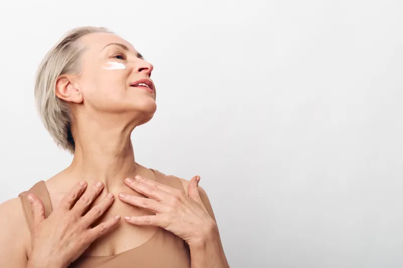 Mature woman with short blonde hair applies white cream on face and neck. Skin care concept with natural beauty and anti aging treatment on neutral background with soft lighting.