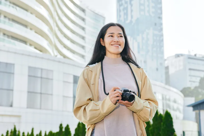 Casual photographer smiling outdoors with camera in hand, urban skyline and modern buildings behind, candid street style and travel vibe for lifestyle, exploration and creative storytelling